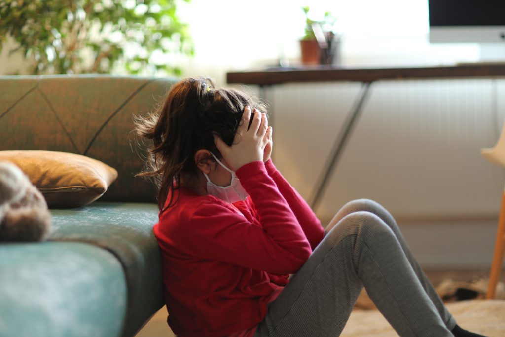 A young person with their hands covering their face leaning against a couch