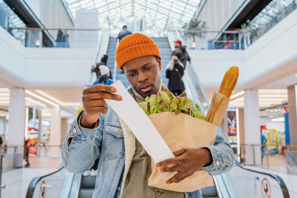 Surprised African-American man in denim jacket looks at receipt total in sales check holding paper bag with products in mall.