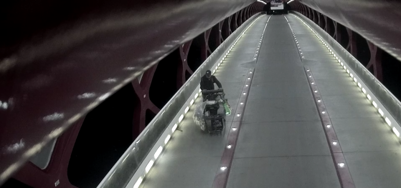 A person pushing a shopping cart along a long bridge is seen via CCTV camera footage