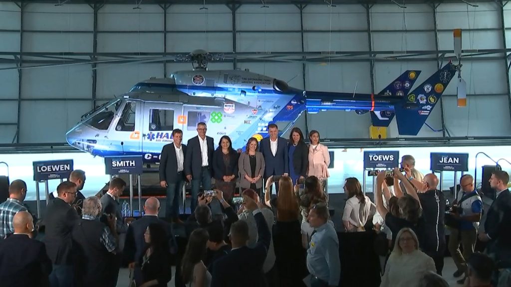 Seven UCP leadership candidates pose for a photo on a stage in front of a row of podiums, with a helicopter in the background.