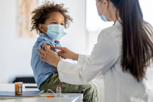 A young child wearing a facemask sits on a doctor's table while a physician prepares to administer a vaccine in their arm