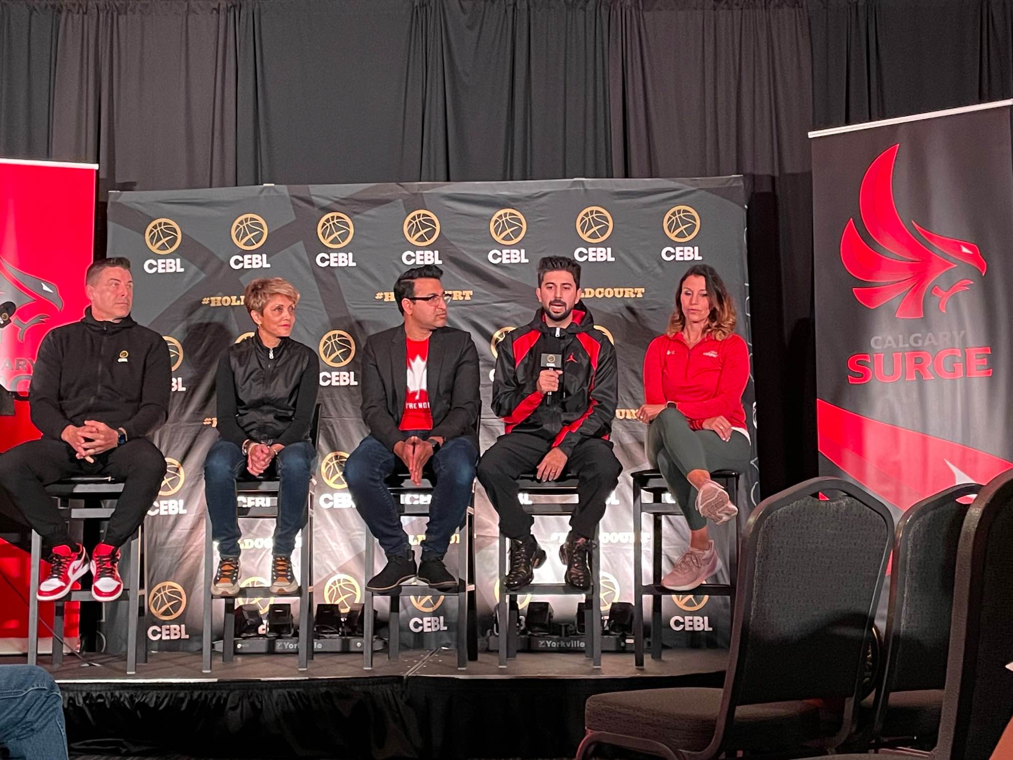 Officials sit on stage to unveil the new Calgary Surge CEBL team name and logo. Red and black banners are draped on either side of the stage bearing a logo design modeled after a bird of prey