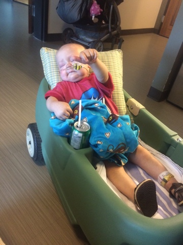 An infant with a nasal cannula lays in a plastic wagon giving a thumbs up while cradling a blanket