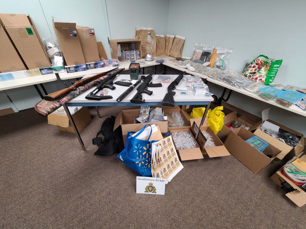 A closer view of seized items including cannabis cigarette packaging, vape pens, printed labels, seized firearms, boxes of illegal cigarettes, pre made illegal cannabis cigarettes, packages of illegal edible cannabis products