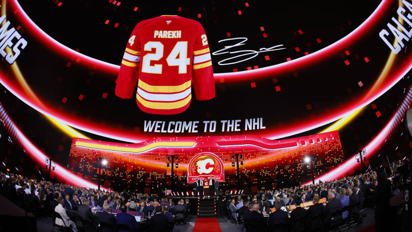 A jersey of Zayne Parekh, the Calgary Flames' first round pick in the 2024 NHL Draft, hangs in the forground as he poses with Flames management on stage